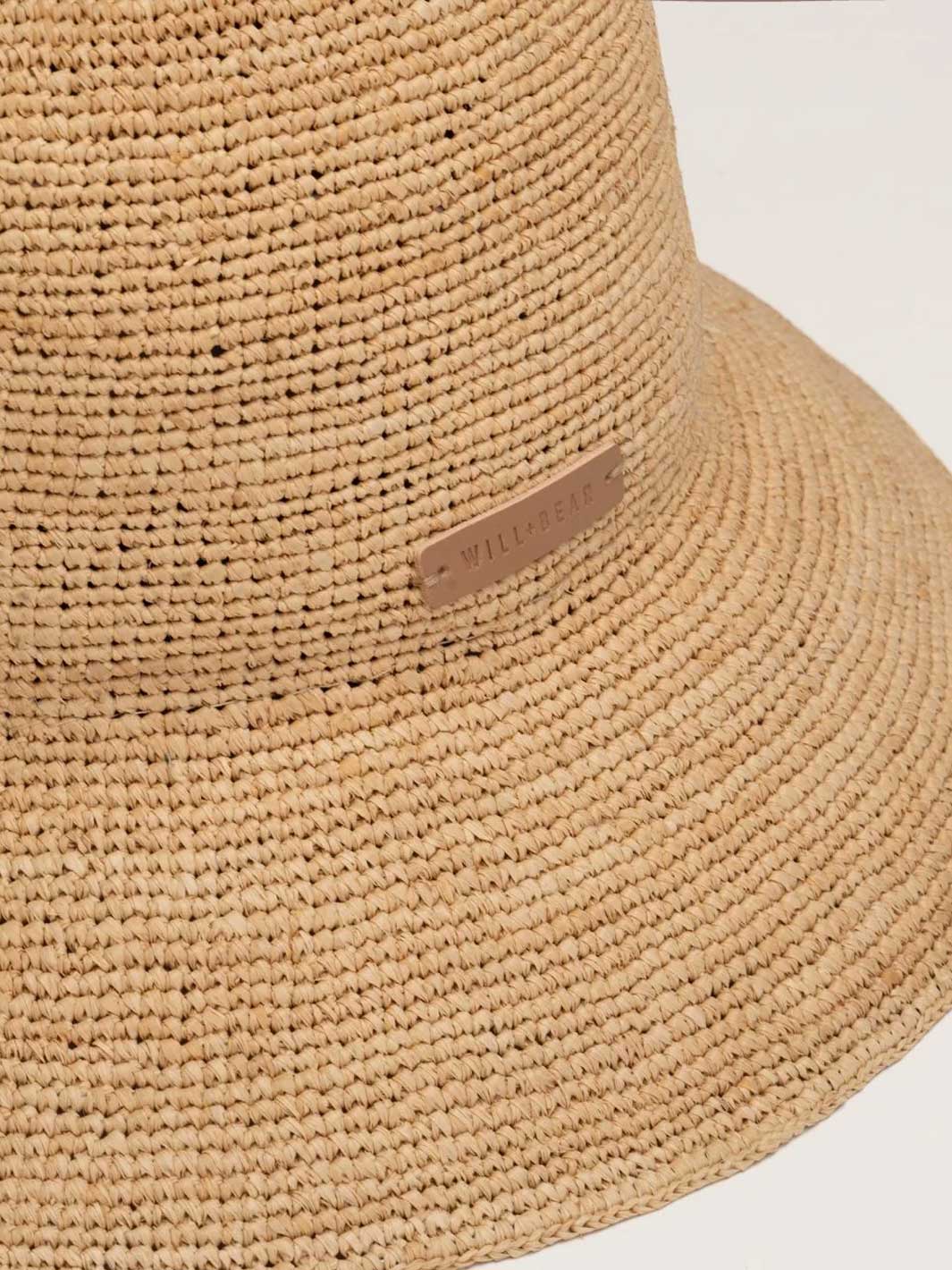 Close-up of a woven straw hat in a natural tan color, showing the textured straw material and a small tan leather brand tag on the crown.