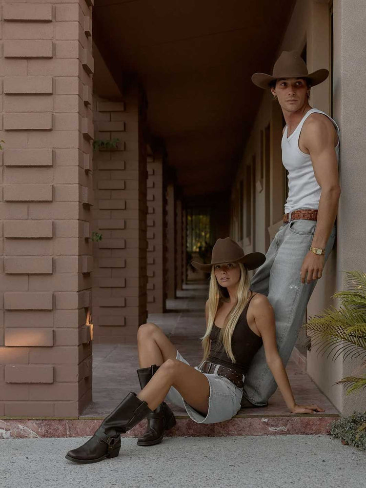 A man and a woman posing in matching brown cowboy hats, tank tops, denim and boots in a covered outdoor walkway.