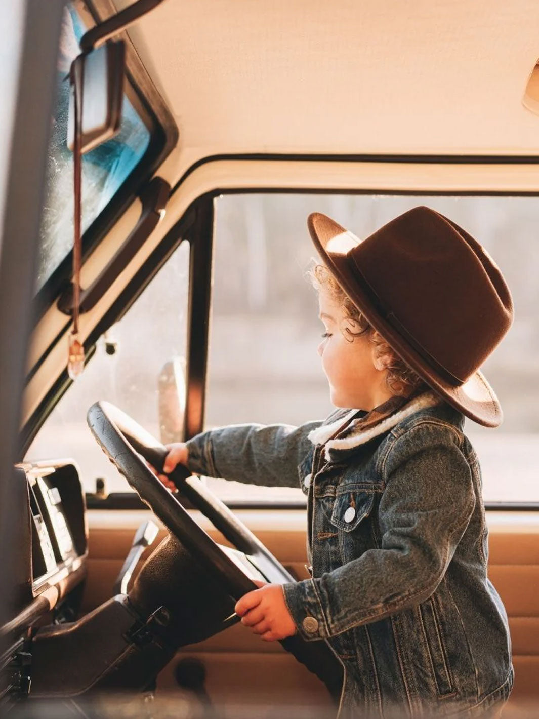 A toddler wearing a brown wide-brim felt fedora hat and a denim jacket sits inside a vintage-style vehicle, holding the steering wheel.