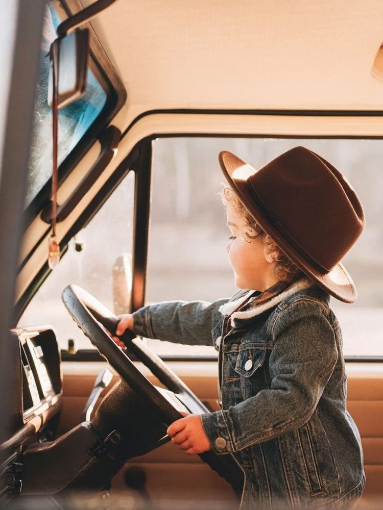 A toddler wearing a brown wide-brim felt fedora hat and a denim jacket sits inside a vintage-style vehicle, holding the steering wheel.