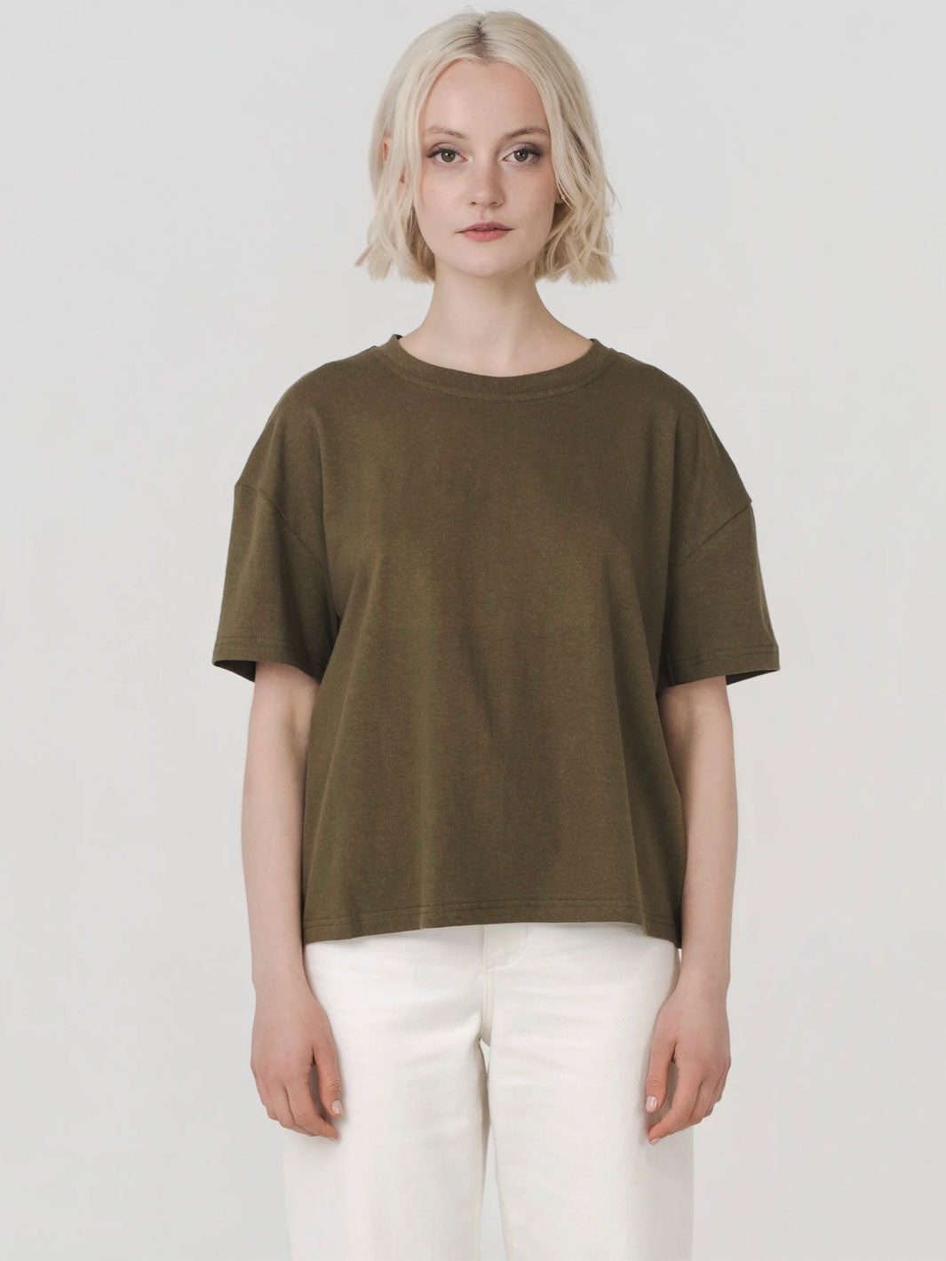 A woman with short blonde hair wearing an oversized olive green short-sleeve t-shirt and white pants, standing against a light gray studio background.