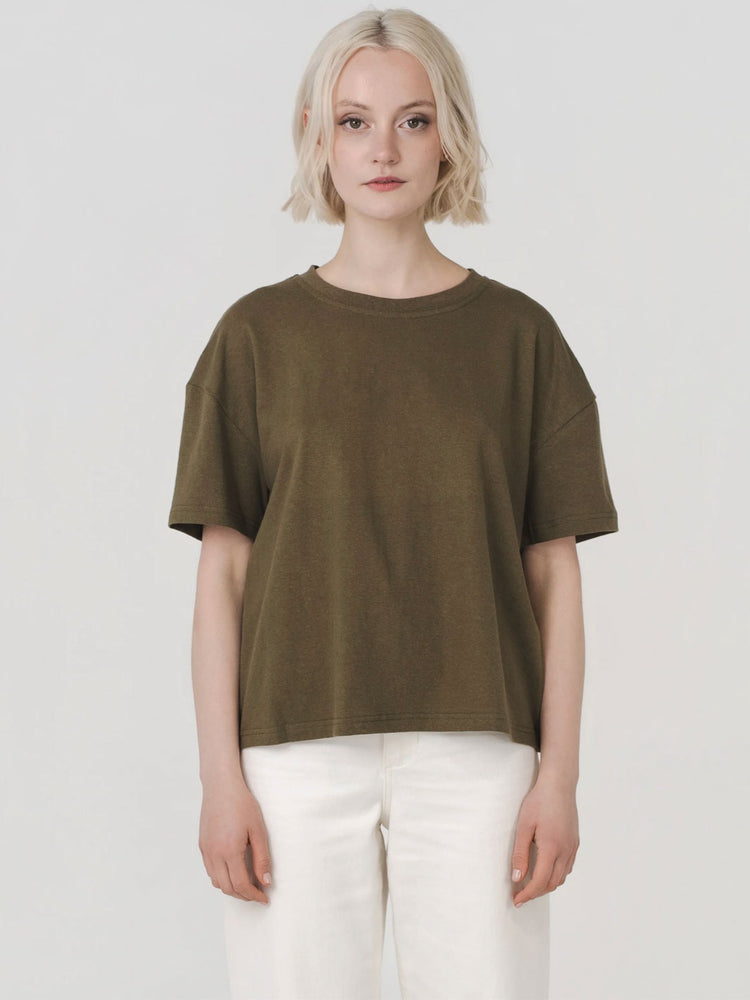 A woman with short blonde hair wearing an oversized olive green short-sleeve t-shirt and white pants, standing against a light gray studio background.